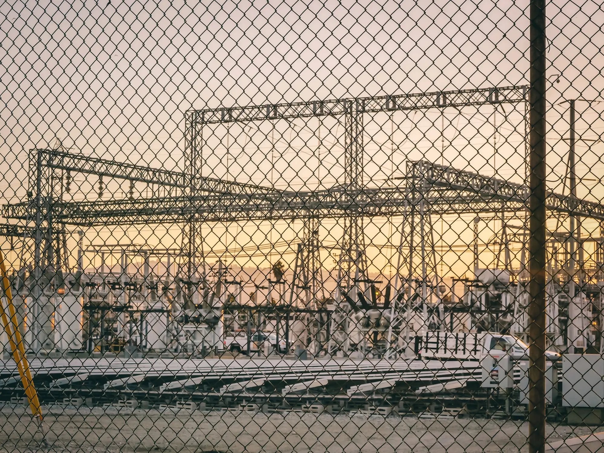Gantries and equipment of a switchyard at dusk, seen through a chain-link fence